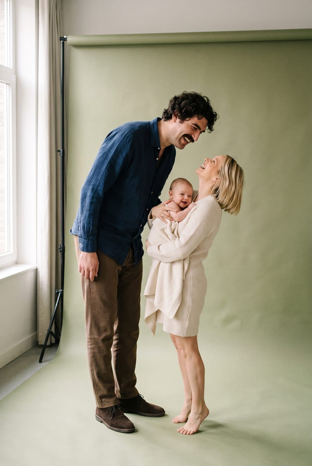 Un père et une mère blonds penchés vers leur petite fille, fond vert sauge